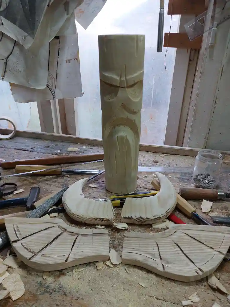 A wooden tiki god carving stands upright on a cluttered workbench surrounded by wood shavings, carving tools, and partially…