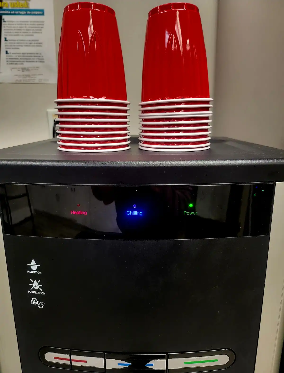 Two stacks of red plastic cups sit atop a black water cooler. The cooler's display shows 'Heating' in red, 'Chilling' in…