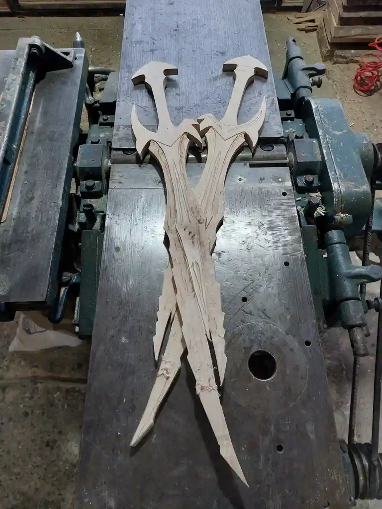 Two intricately carved wooden sword-like objects with ornate, fantasy-style hilts and blades lie on a metal workbench of a…