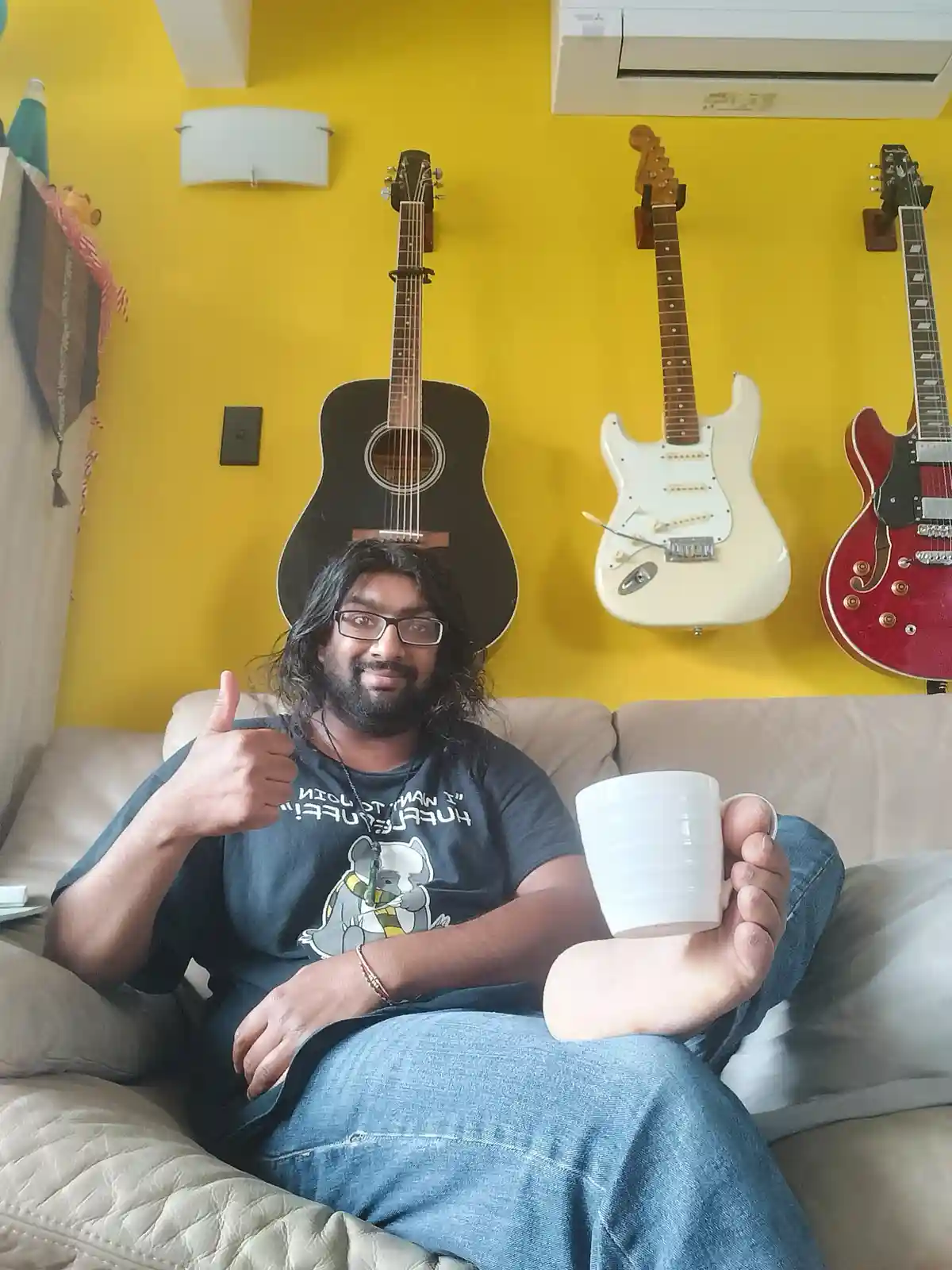 A man with long hair and glasses sits on a beige couch, giving a thumbs-up while holding a white mug with his foot. Behind…