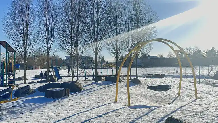 A snowy playground with bare trees and bright sun, featuring swings, slides, and climbing structures, with a few people in…