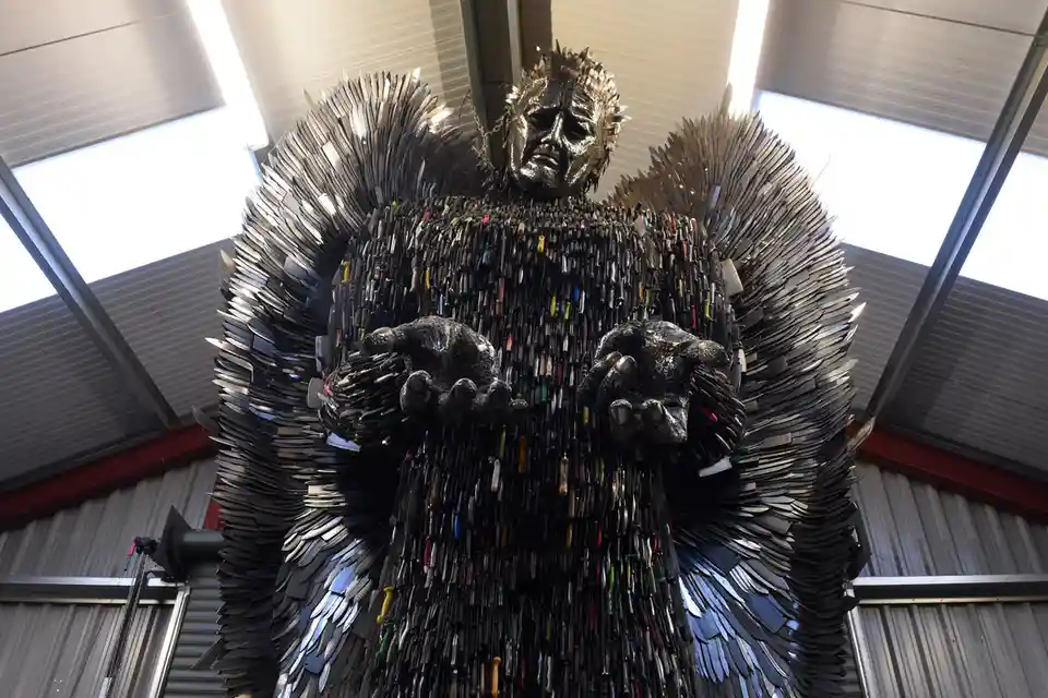 A towering sculpture of a humanoid figure with wings and a distressed face, entirely constructed from thousands of knives…