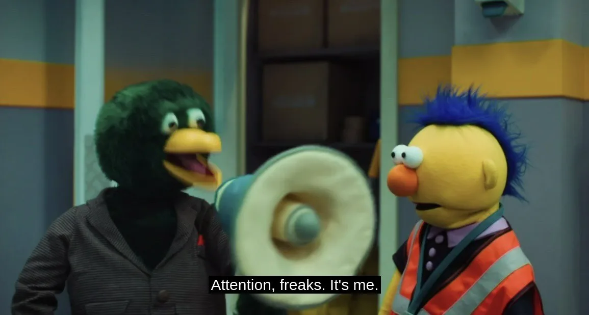 A green duck puppet in a gray jacket holds a megaphone and shouts at a yellow puppet with blue spiky hair wearing a safety…