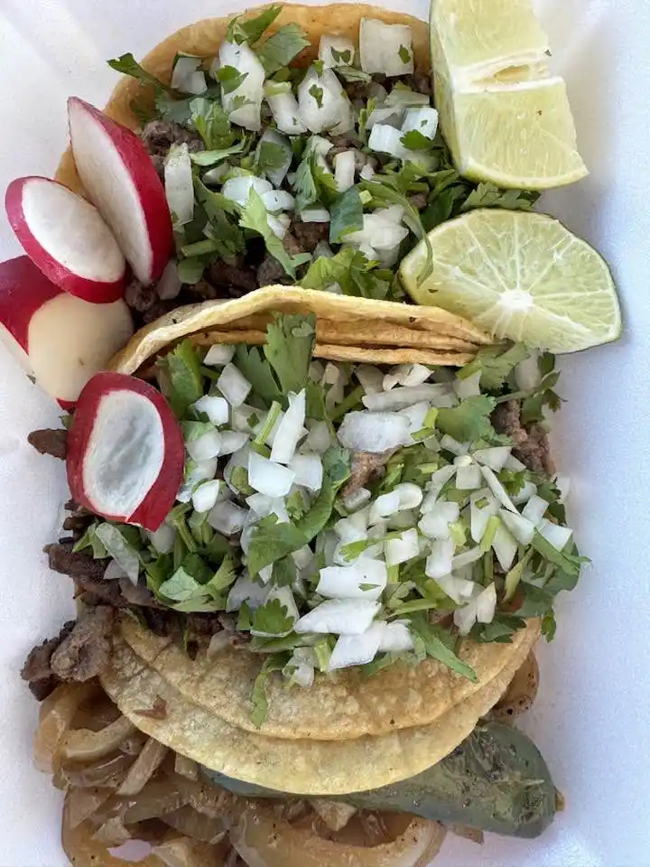A close-up photo of a takeout container holding several tacos topped with chopped cilantro, onions, and meat, accompanied by…