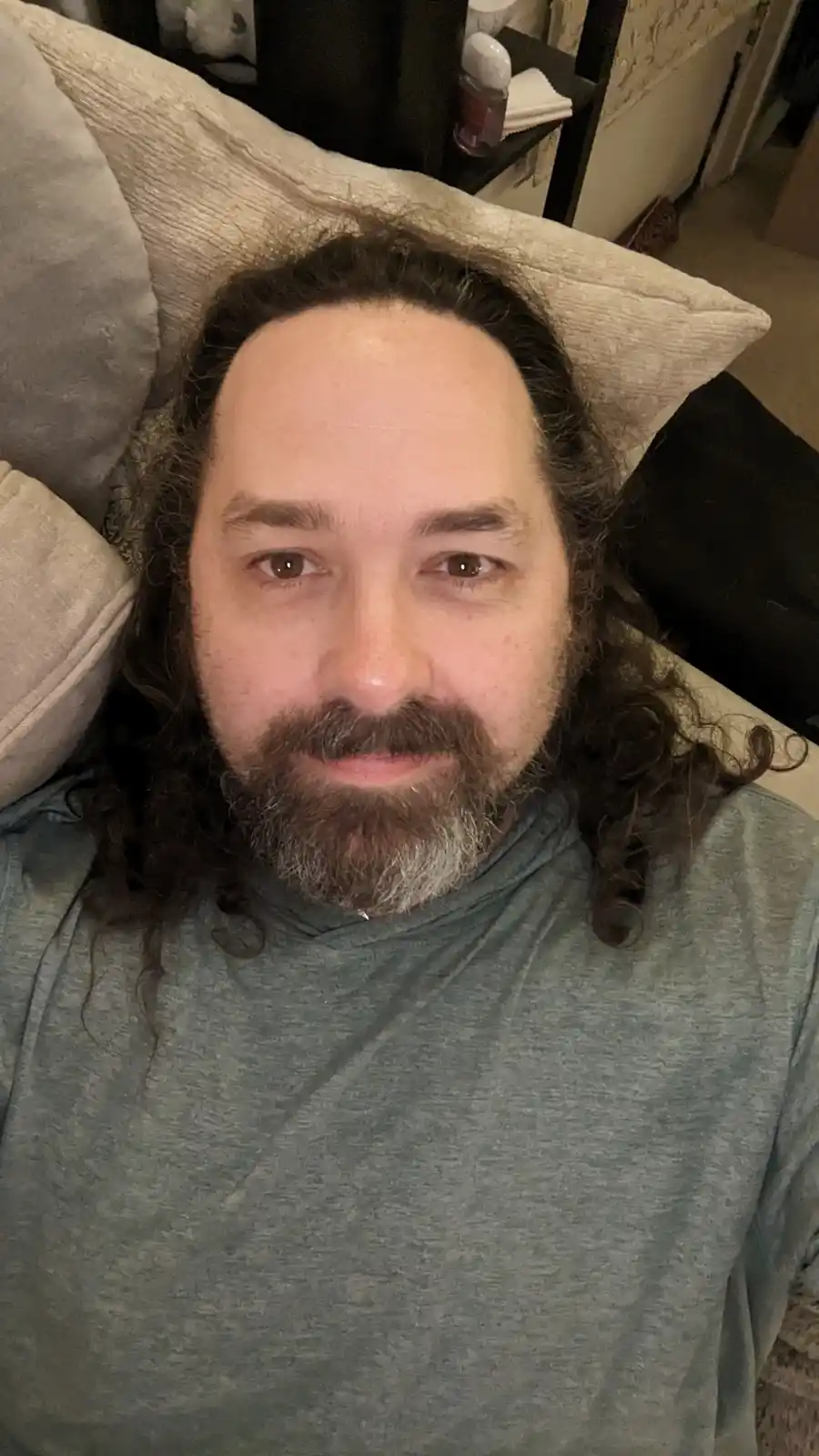 A man with long dark curly hair and a full beard, wearing a gray hoodie, lies on a couch with pillows behind his head. He…