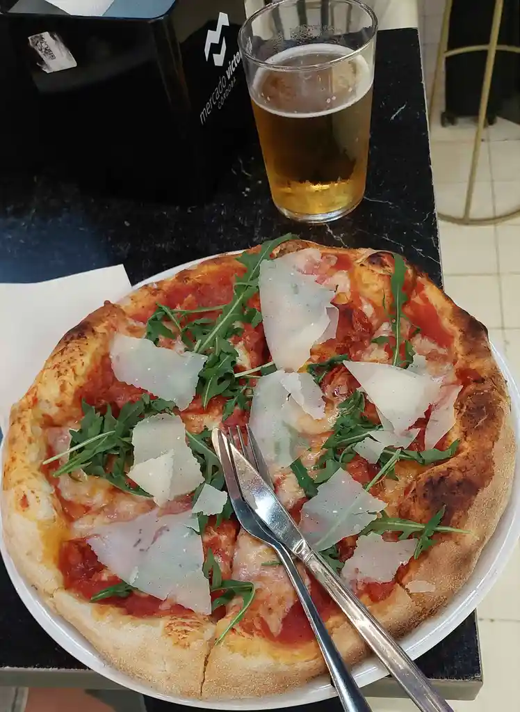 A close-up photo shows a freshly baked pizza on a white plate, topped with tomato sauce, melted cheese, arugula, and shaved…