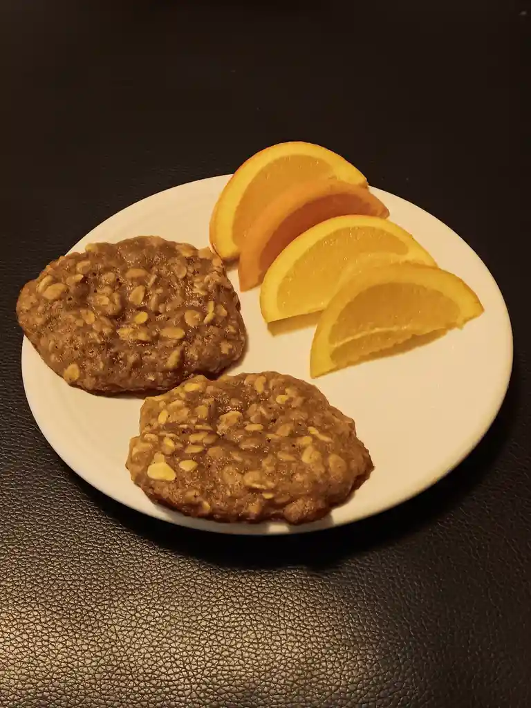 A white plate holds two oatmeal cookies and four orange slices arranged on a dark textured surface. The overlaid text reads…