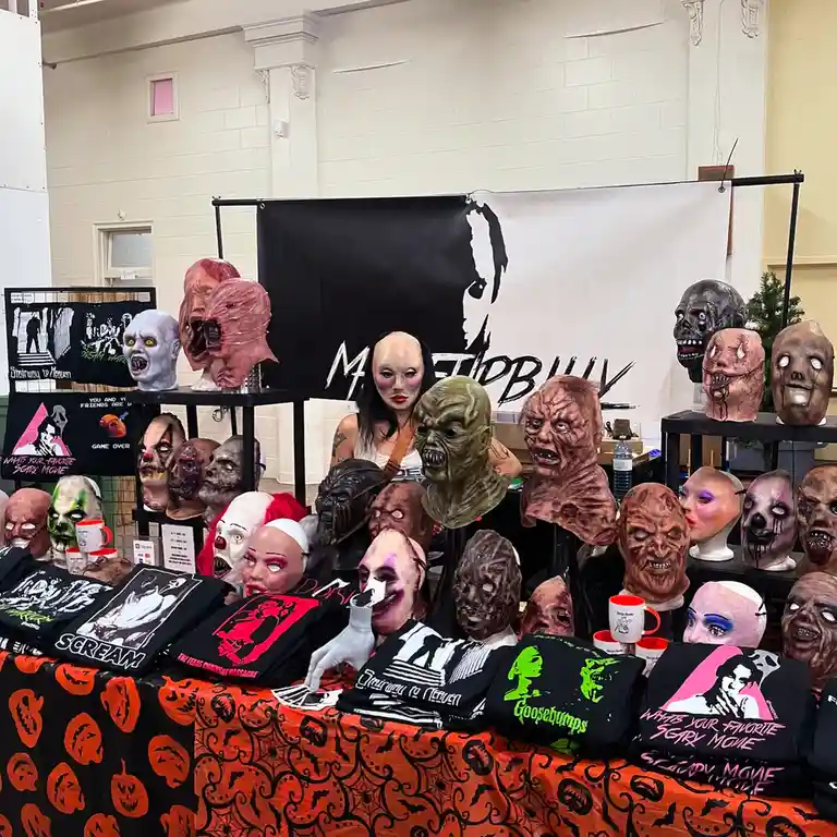 A vendor booth at a convention displays numerous horror movie masks and themed t-shirts on an orange tablecloth with black…
