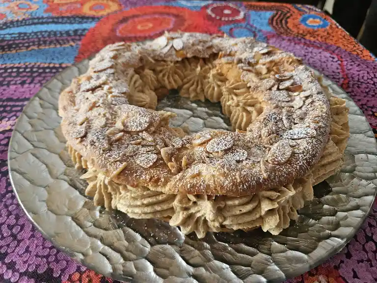 A ring-shaped cake with piped cream filling, sliced almonds, and powdered sugar sits on a textured glass plate atop a…