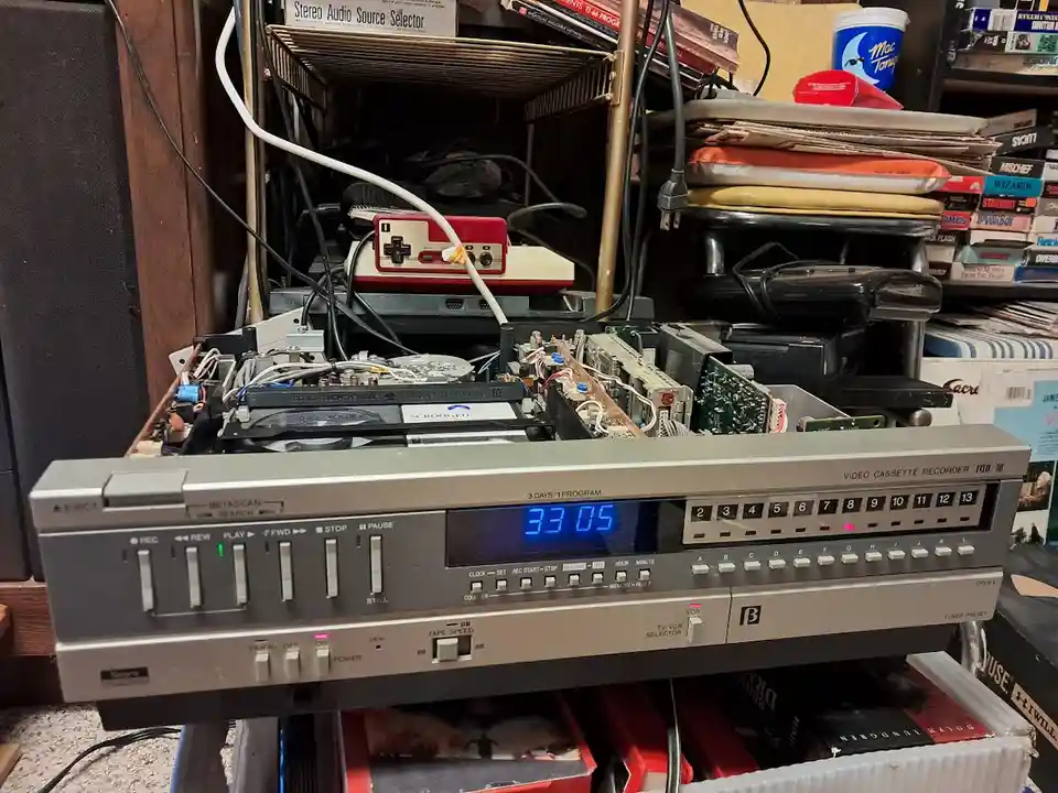 A silver video cassette recorder (VCR) with its internal circuitry exposed and wires dangling, sits in a cluttered room…