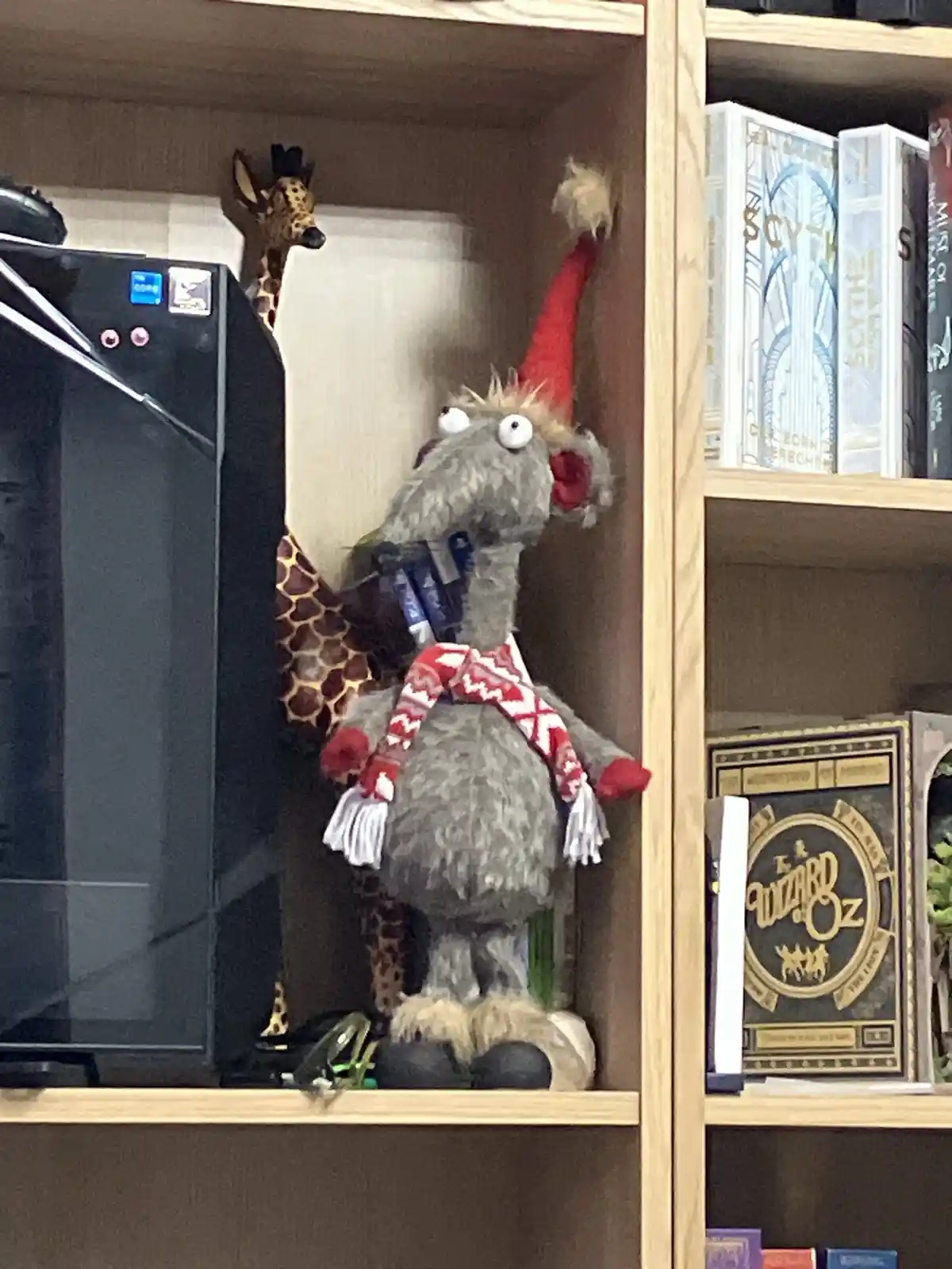 A grey, furry stuffed llama wearing a red pointed hat and a red-and-white patterned scarf is positioned on a wooden shelf…