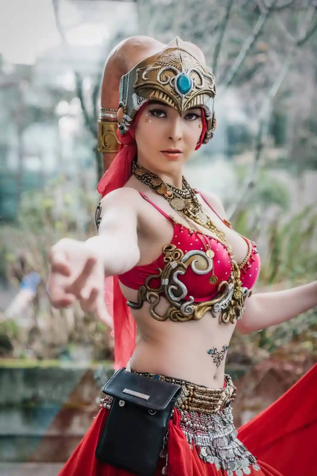 A woman in elaborate fantasy cosplay, wearing a red outfit with ornate gold and silver armor pieces, a helmet with a blue…