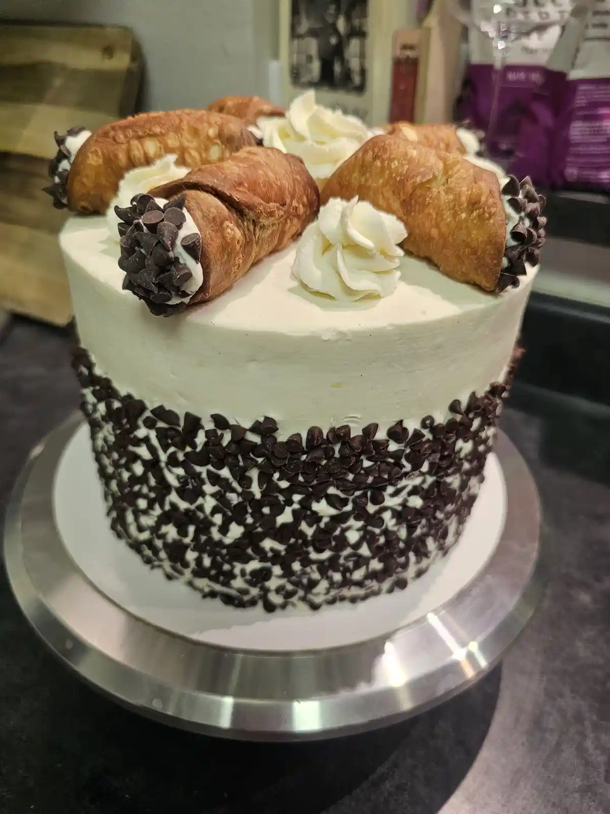 A white-frosted cake decorated with chocolate chips around its base and topped with several golden-brown cannoli shells…
