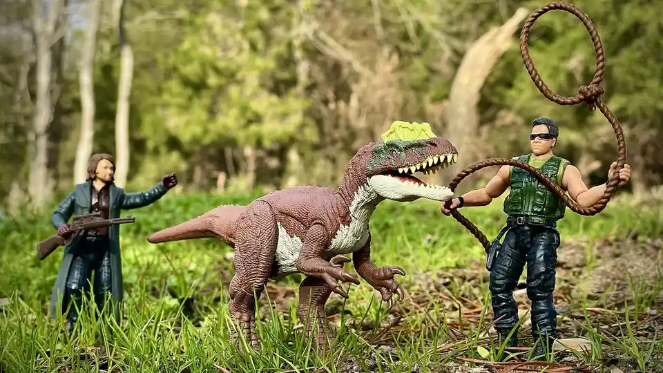 A toy action figure resembling a cowboy with a lasso is positioned to rope a toy dinosaur in a grassy outdoor setting…