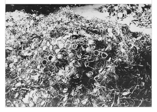 a black-and-white historical photograph shows a massive pile of discarded eyeglasses and frames, mixed with debris, filling…