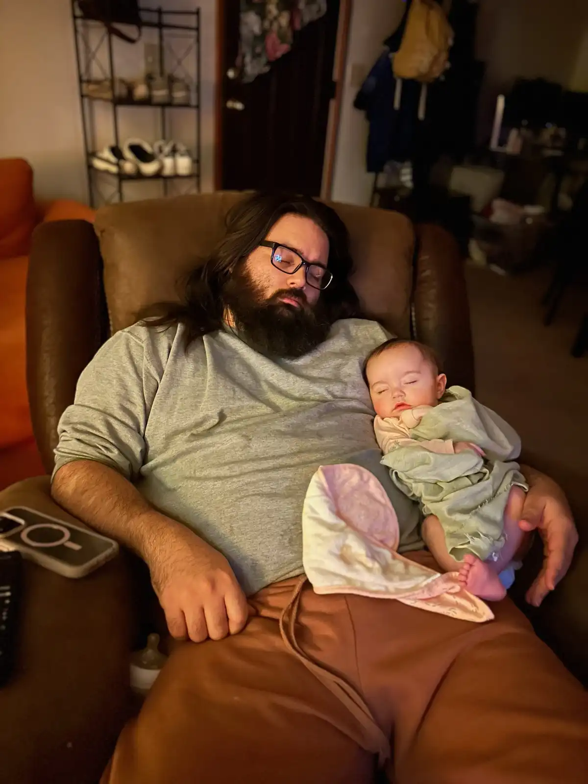 A man with long hair and a beard, wearing glasses and a grey shirt, is asleep in a brown recliner while holding a sleeping…
