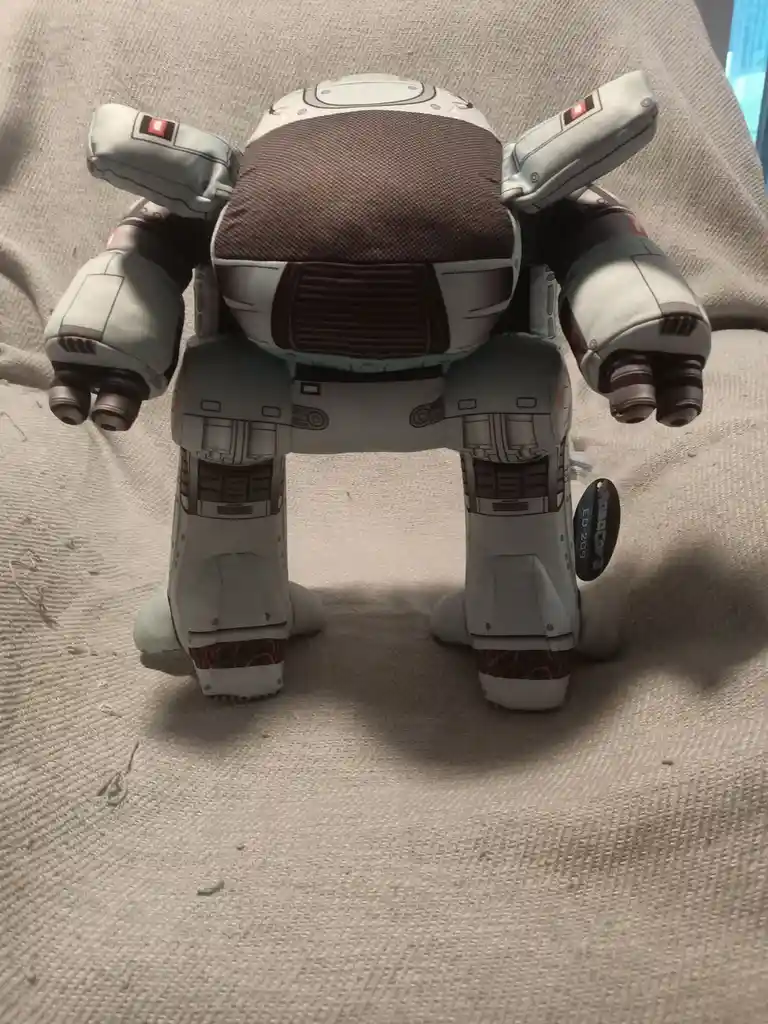 A plush toy of a bipedal robot, viewed from behind, stands on a textured beige fabric surface. The robot has a bulky, light…