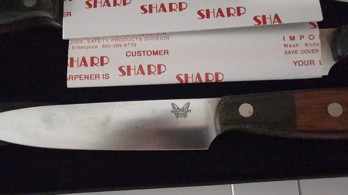 A close-up photo shows a Benchmade knife with a wooden handle resting on a dark surface, positioned beneath a white paper…