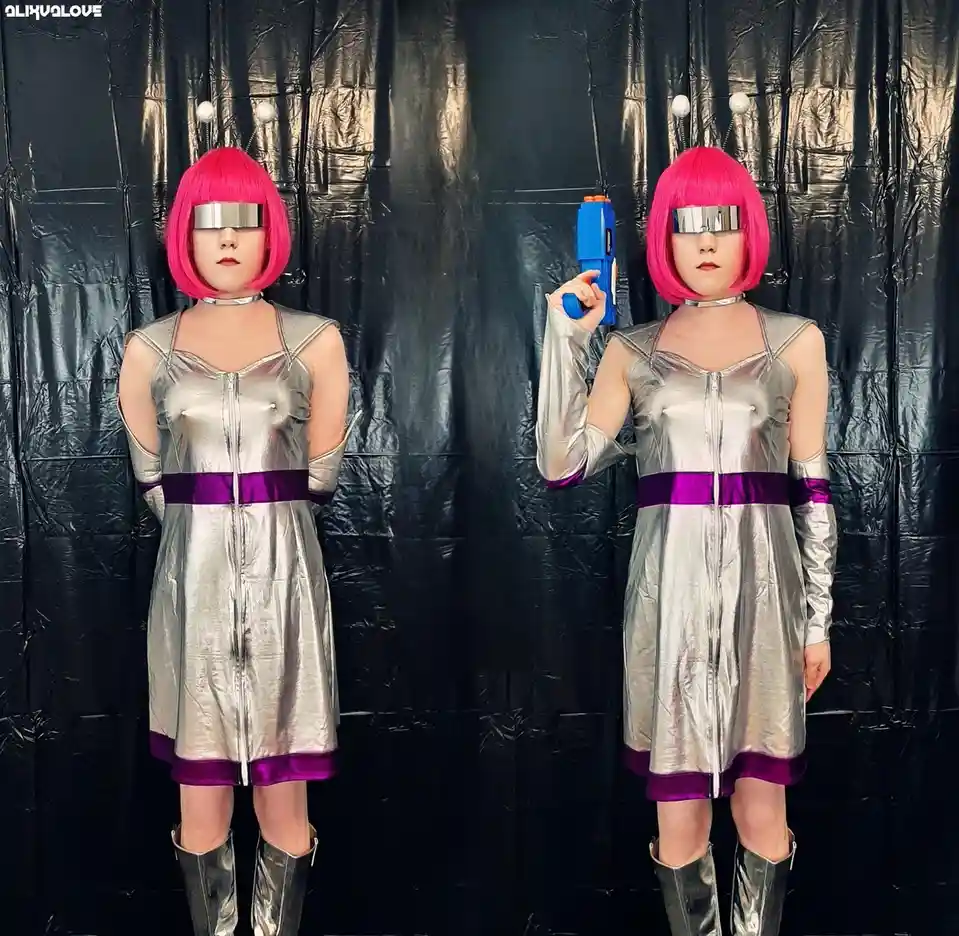Two people dressed as identical alien characters with pink bob wigs, silver metallic outfits with purple trim, and antennae…
