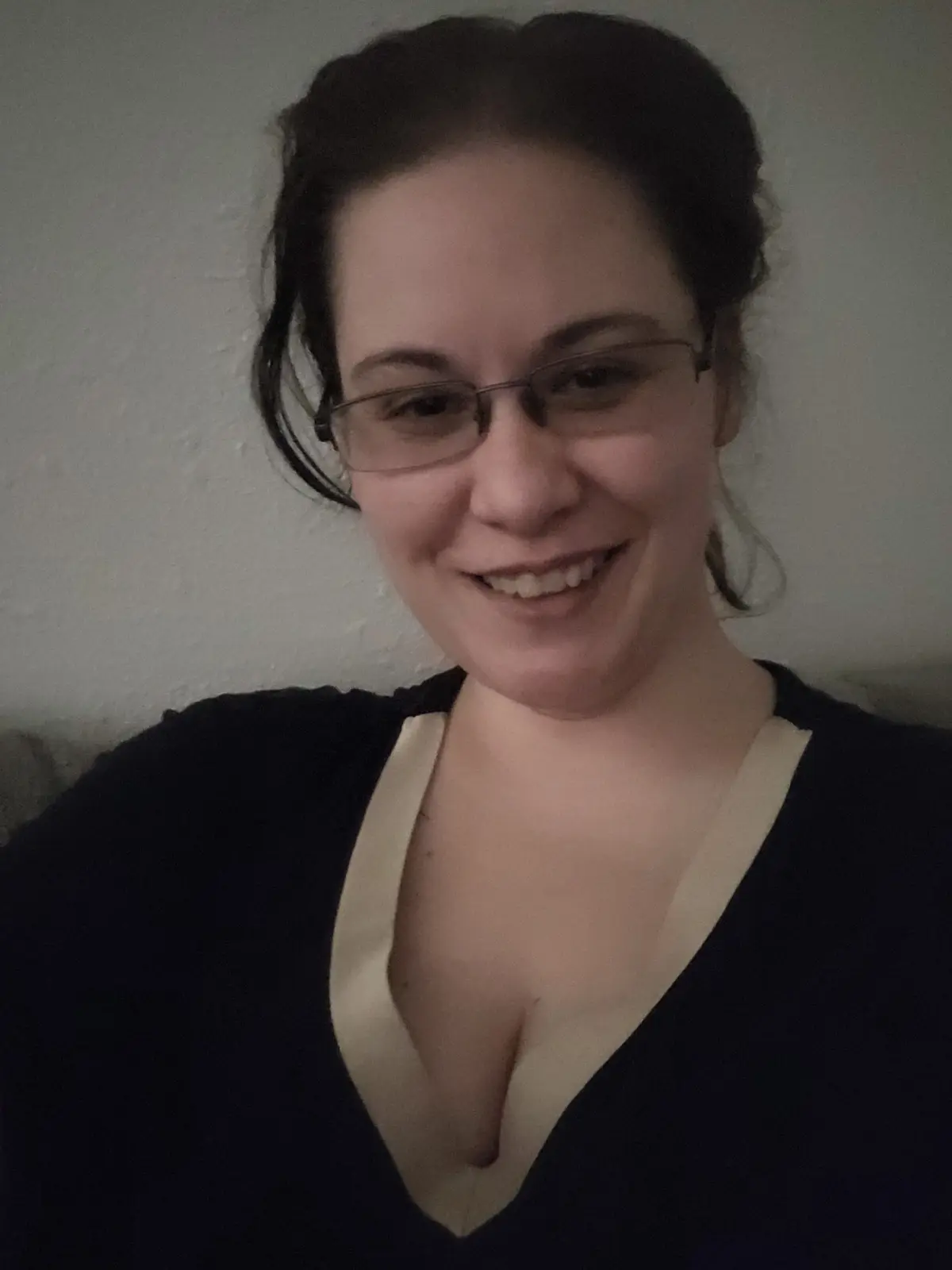 A woman with dark hair pulled back and wearing glasses smiles at the camera while wearing a dark v-neck shirt with a…