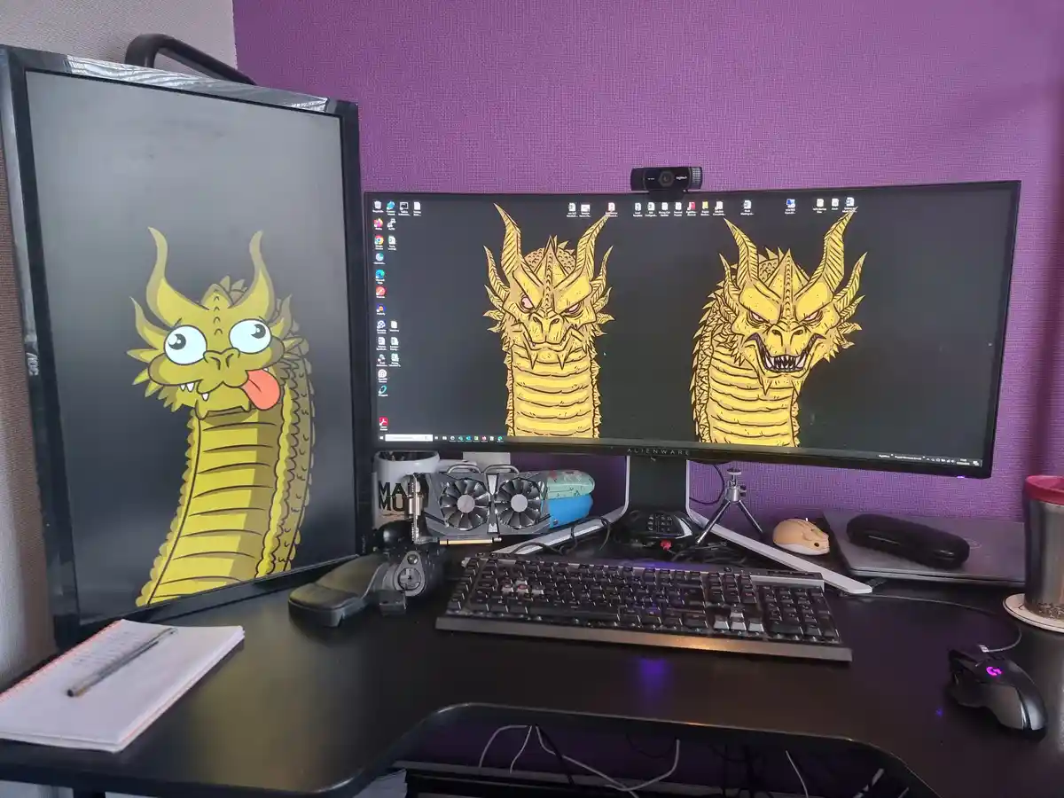 A computer desk setup with two monitors; the left monitor shows a cartoonish yellow dragon with a goofy expression, while…