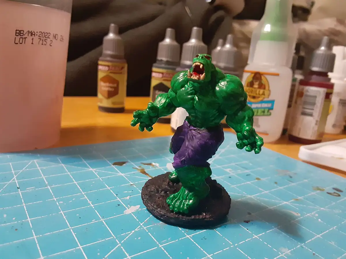 A painted green Hulk figurine with purple pants stands on a blue gridded mat, surrounded by craft supplies like paint…
