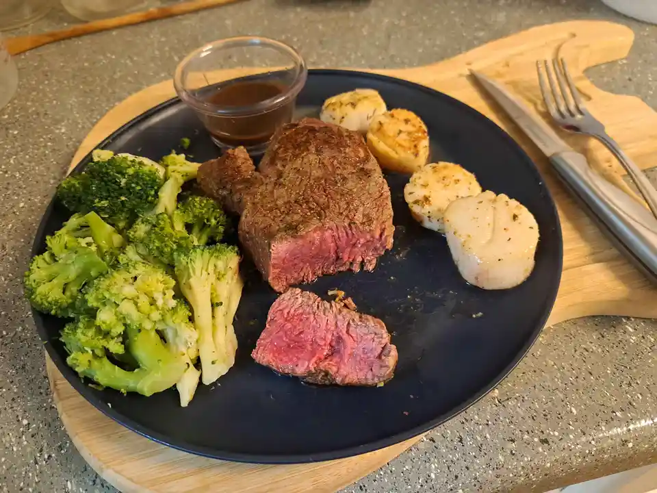 A plate of food featuring a medium-rare steak with a slice cut off, showing pink interior, accompanied by roasted scallops…