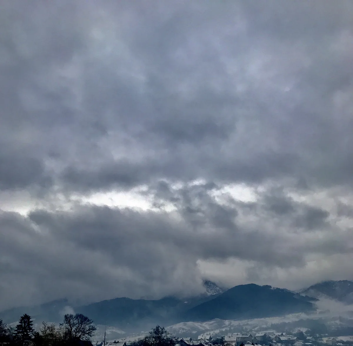 A moody, overcast sky with heavy gray clouds dominates the image, partially obscuring snow-dusted mountains and a small…