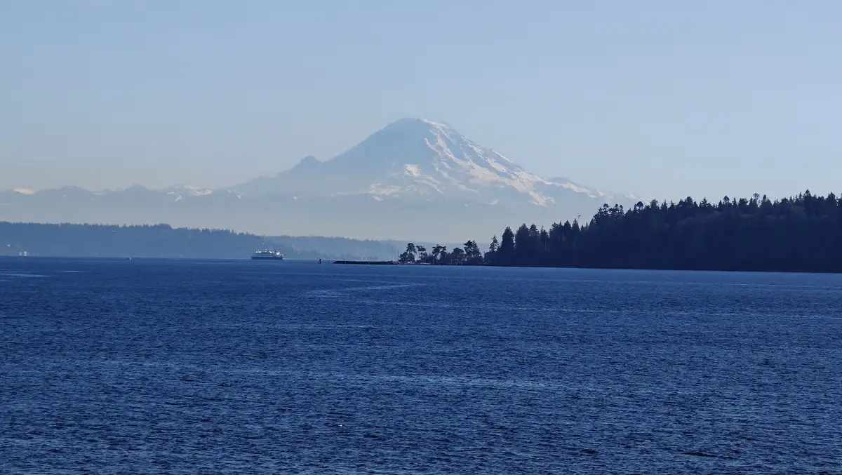 A scenic view of a large body of water with a snow-capped mountain in the distance under a hazy sky. A ferry is visible on…