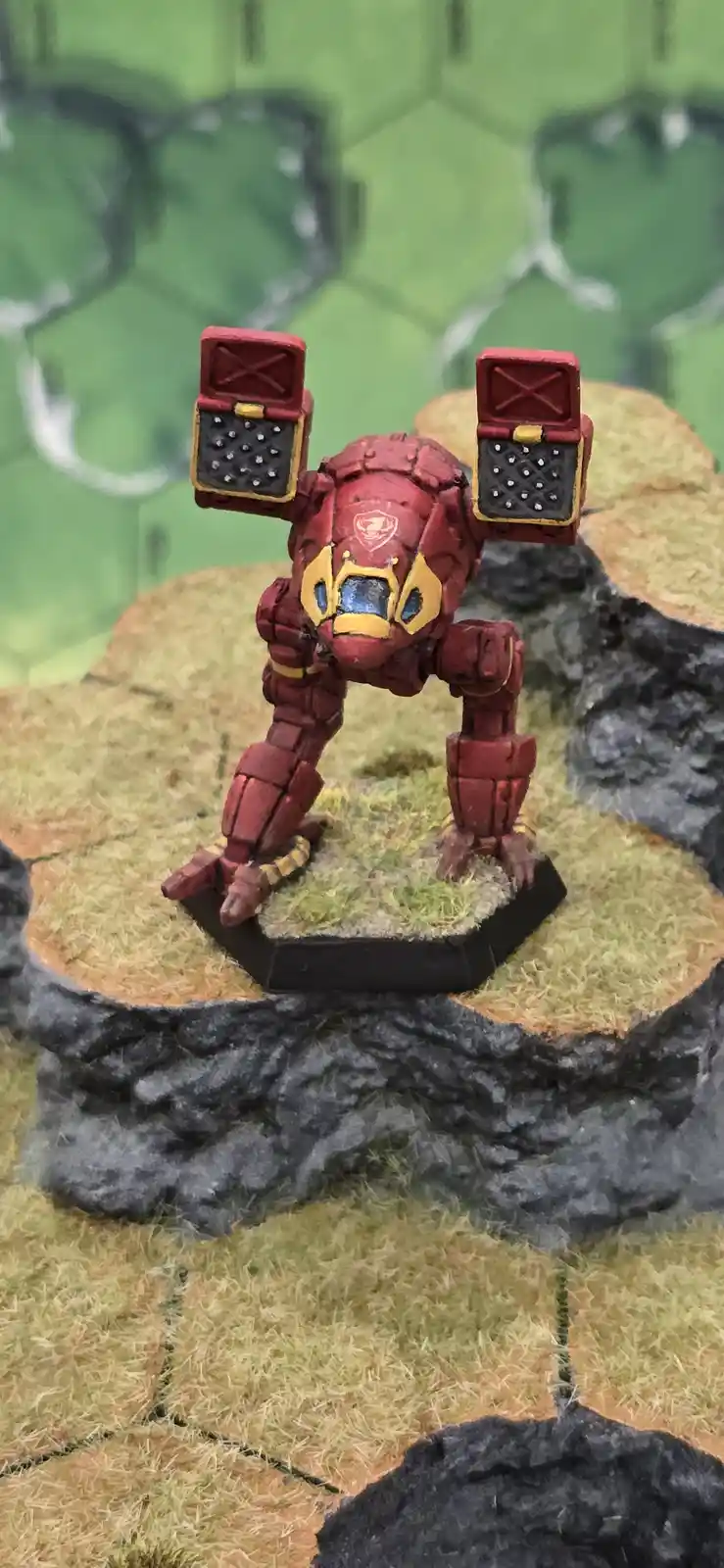 A red and gold painted miniature robot figurine stands on a grassy terrain tile, its arms raised with open hatches revealing…