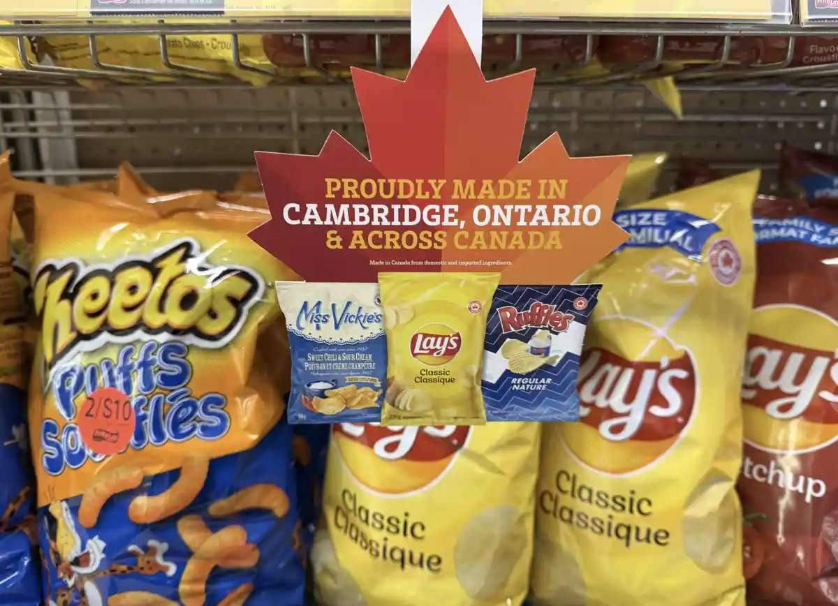 A supermarket shelf displays bags of Lay's, Ruffles, and Cheetos chips, with a red maple leaf-shaped sign hanging above…