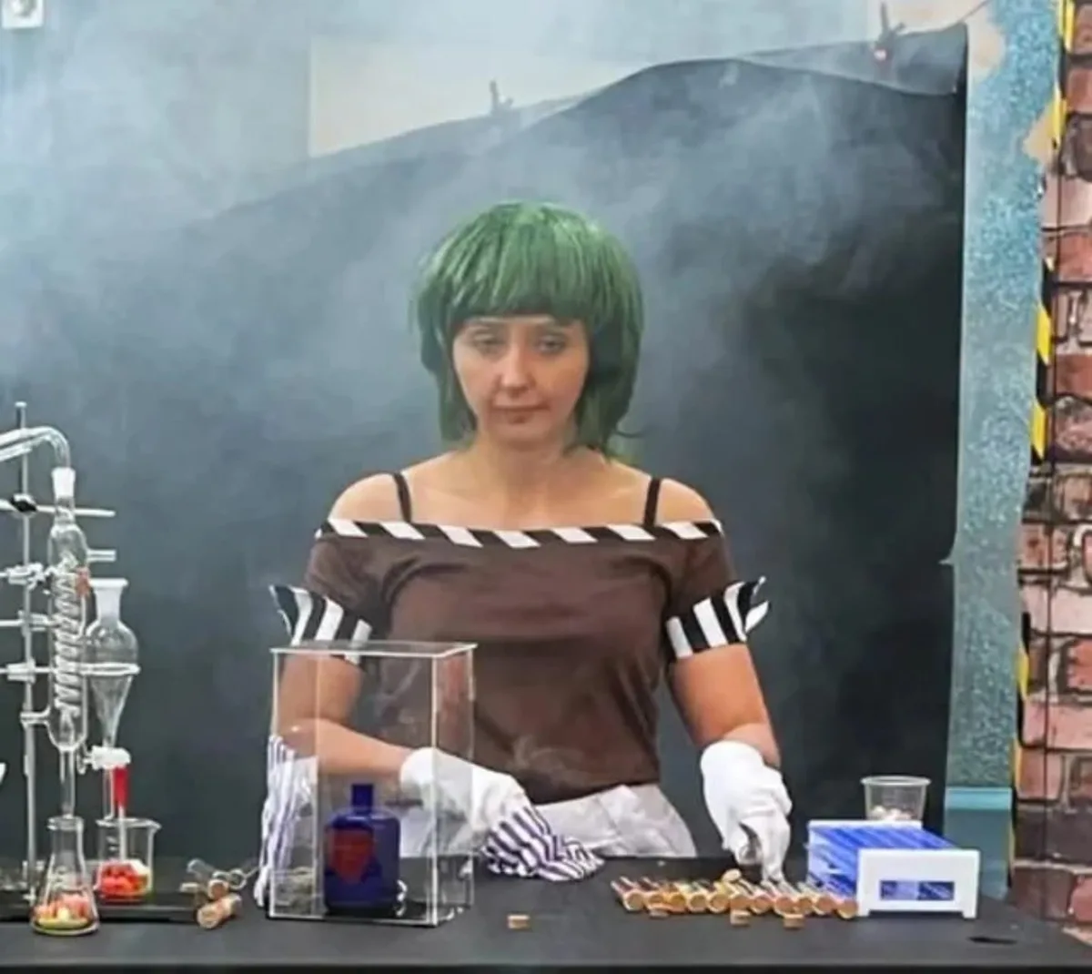 A woman with a green wig and white gloves stands behind a lab table filled with glassware, a blue bottle, and scattered…
