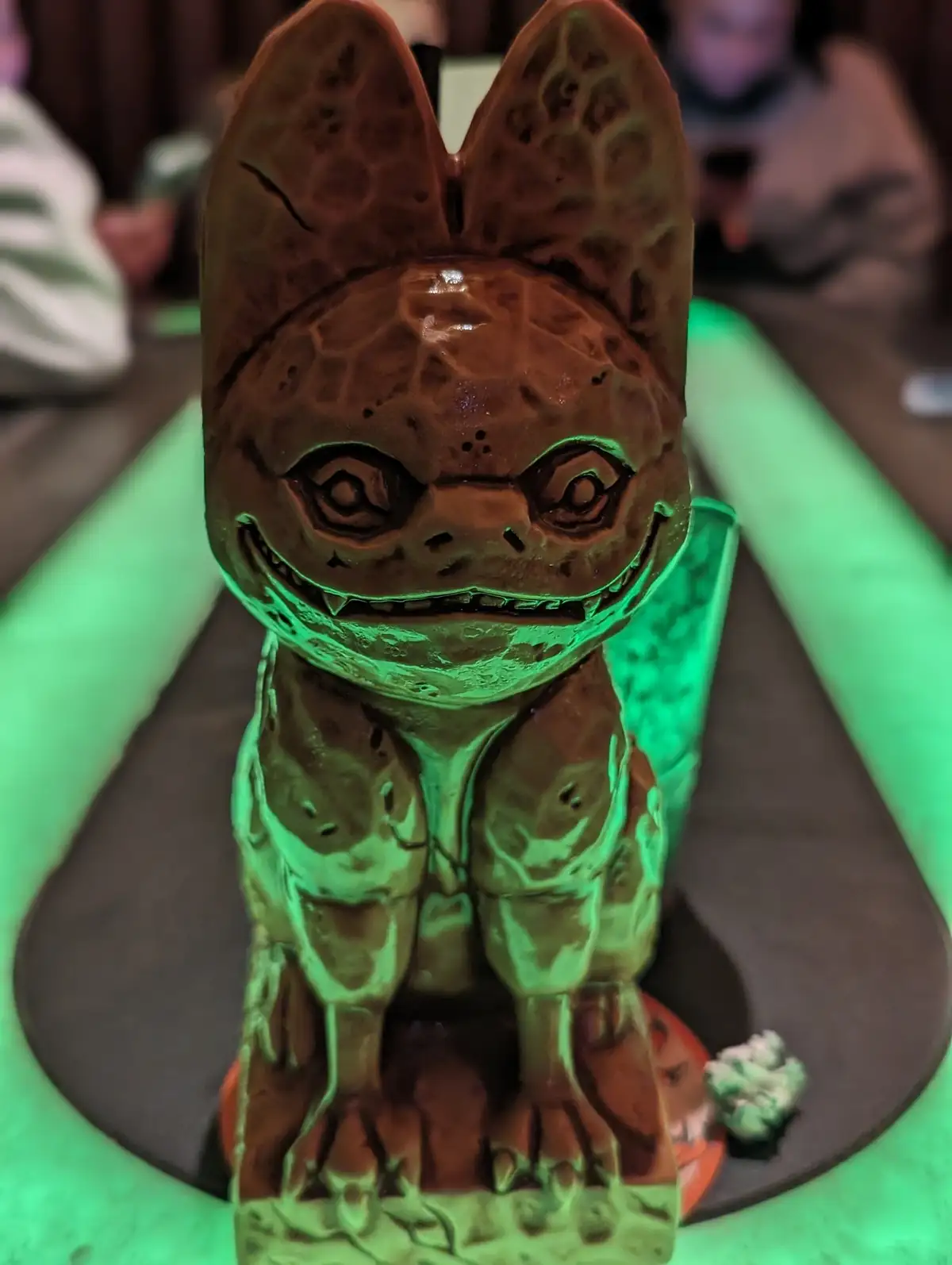 a close-up photo shows a bat-shaped chocolate dessert with large ears, wide eyes, and a toothy grin, illuminated by green…