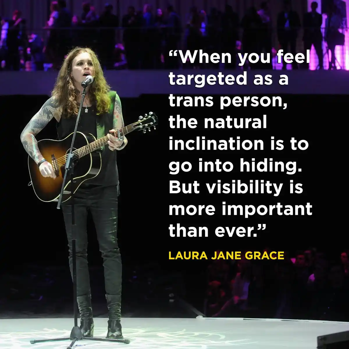 A person with long hair and tattoos stands on a stage holding an acoustic guitar and singing into a microphone. The image…