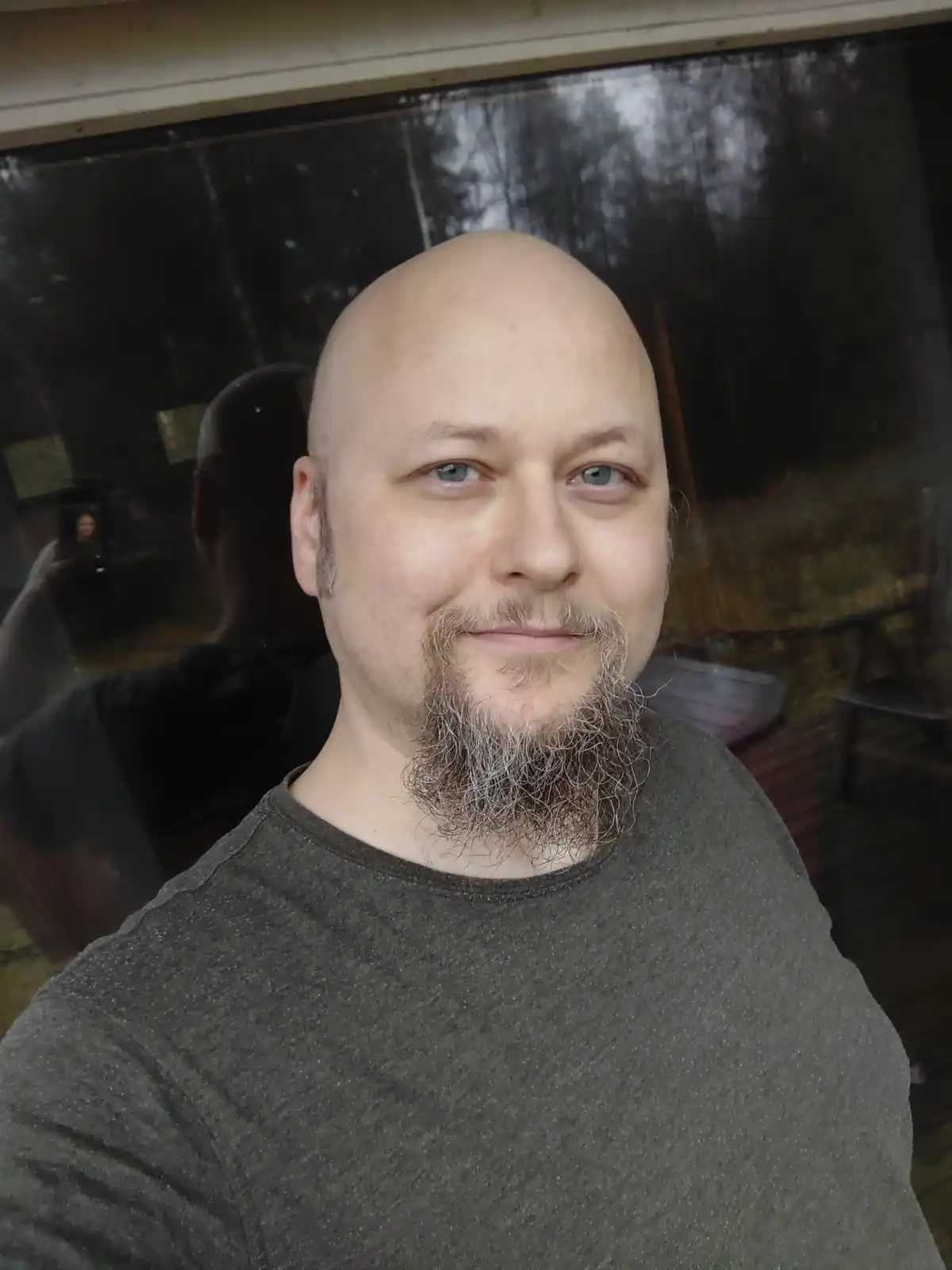 A bald man with a full, graying beard and mustache smiles at the camera while taking a selfie. His reflection is visible in…