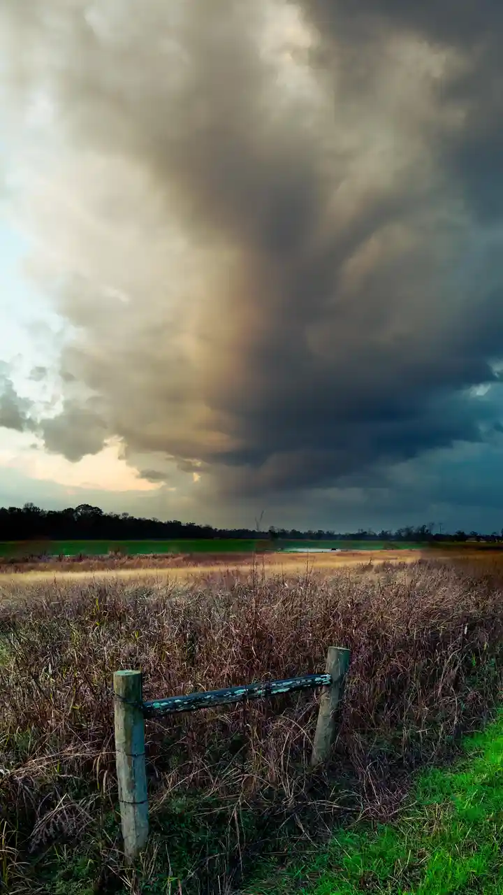 A dramatic landscape photo shows a weathered wooden fence gate in a field of tall, dry grass under a massive, dark storm…