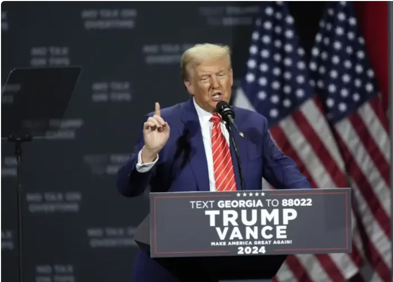 A man in a blue suit and red tie stands at a podium with American flags behind him, pointing upward with his right index…