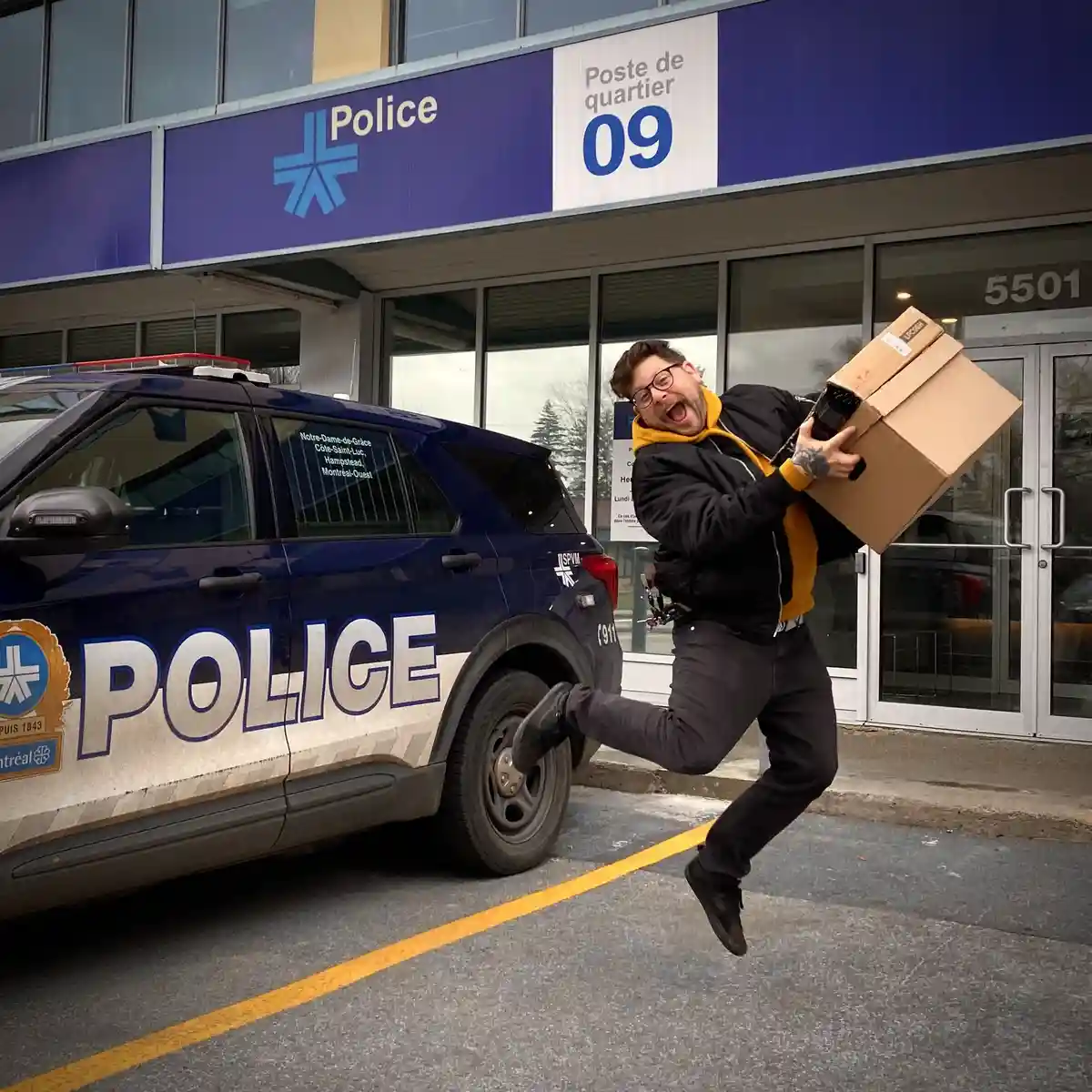 A man in a black jacket and yellow hoodie is mid-air, jumping joyfully while holding a cardboard box, in front of a blue…