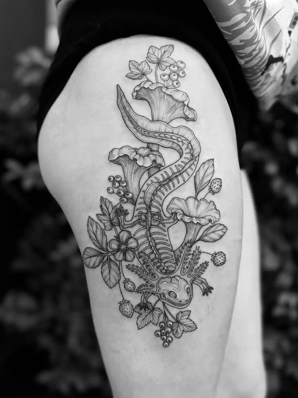 A black and white photo shows a detailed tattoo on a person's thigh, featuring a skeletal newt or salamander intertwined…