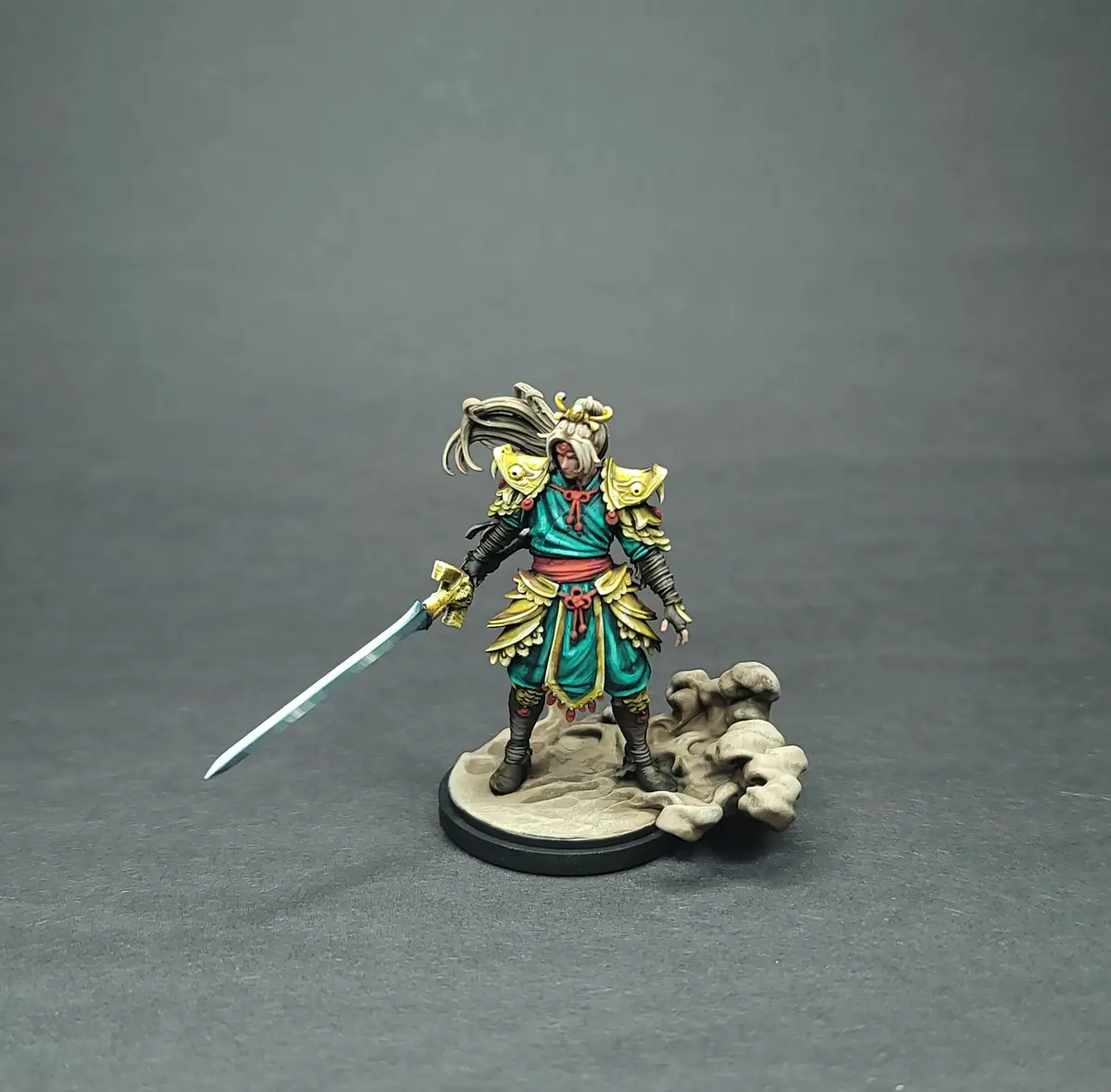 A painted miniature figurine of a samurai-style warrior with long hair, gold armor, and a sword stands on a rocky base…