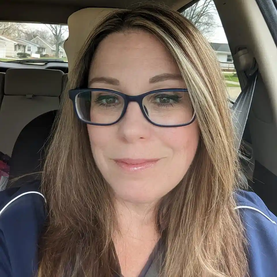 A woman with brown and blonde hair and dark-rimmed glasses is taking a selfie from inside a car, wearing a blue top with…