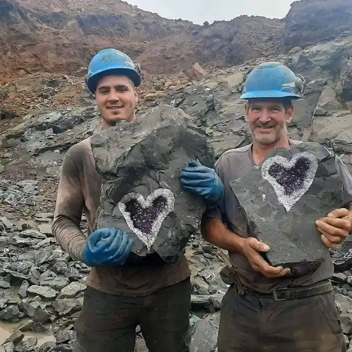 Two smiling men in blue hard hats and work clothes stand in a rocky quarry, each holding a large grey rock with a heart…