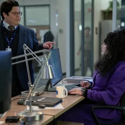 A man in a suit and glasses stands beside a woman in a purple sweater at a desk, pointing at her computer screen. She is…