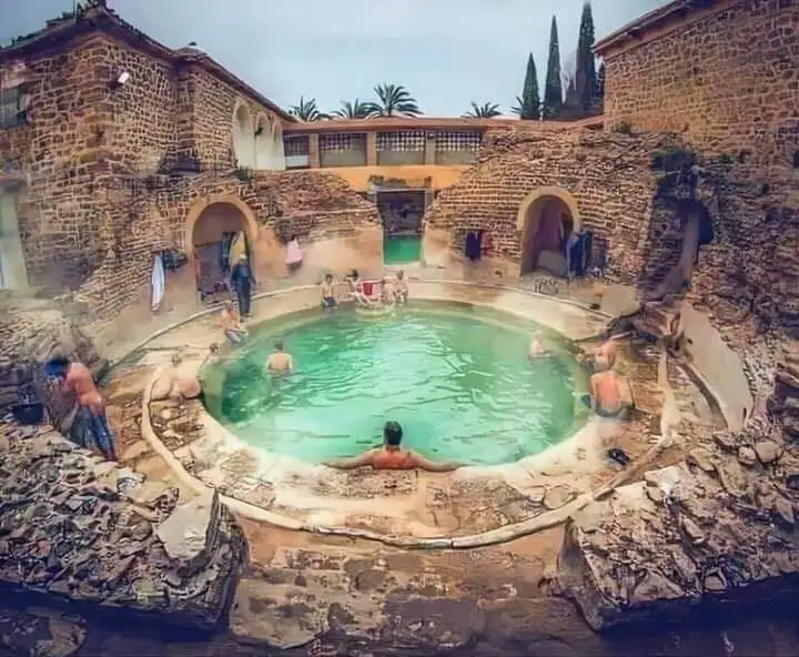 a fisheye lens photo shows people relaxing in a circular, green-tinted pool surrounded by ancient, crumbling stone ruins and…