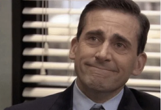a close-up of the character Michael Scott from The Office, wearing a suit and tie, with a forced, awkward smile while…
