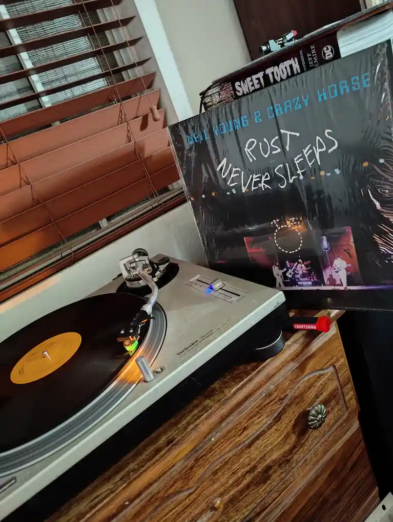 A Technics turntable plays a record with a yellow center label, next to a Neil Young & Crazy Horse album cover that has…
