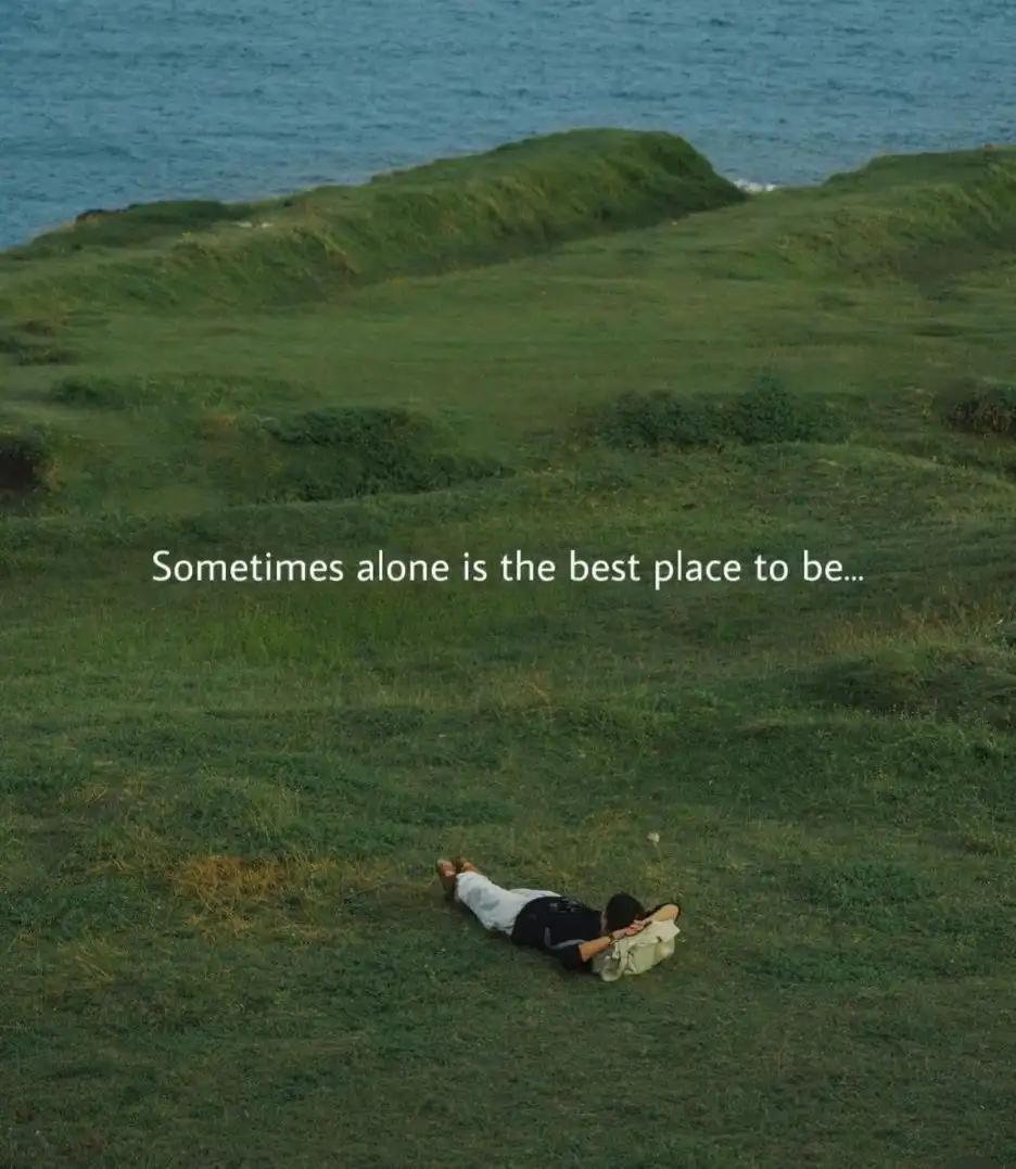 A person lies alone on a grassy hillside overlooking the ocean, with the text 'Sometimes alone is the best place to be...'…