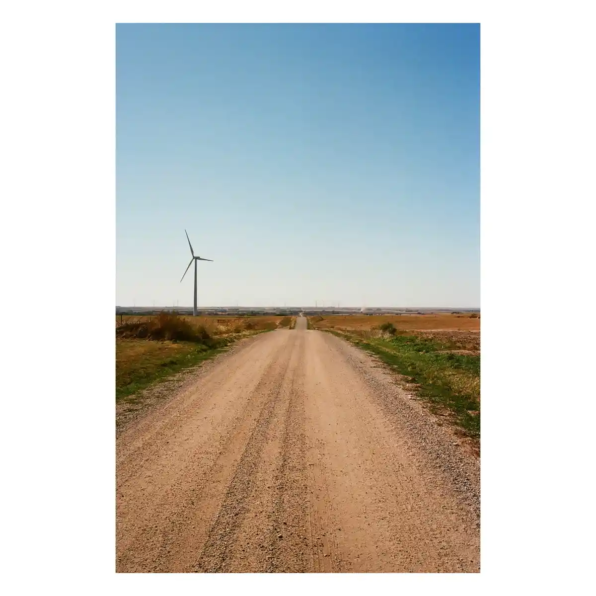 A wide, straight dirt road stretches into the distance under a clear blue sky, flanked by dry grass and fields. A single…