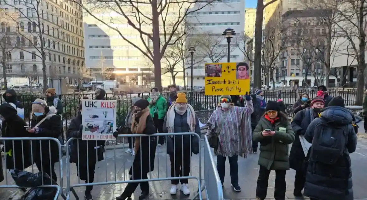 A group of protesters in winter coats and masks stand behind metal barricades in an urban park, holding signs. One sign…