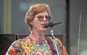 A woman with short reddish hair and sunglasses is singing into a microphone while wearing a colorful floral shirt. The text…